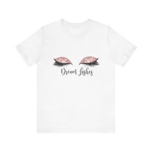May include: White t-shirt featuring a design of closed eyes with pink glitter eyeshadow and long black eyelashes. The words "Dream Lashes" are written in a cursive font below the eyes.
