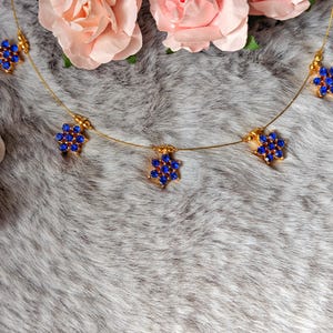 Könnte beinhalten: Eine goldfarbene Halskette mit fünf blumenförmigen Anhängern, die jeweils mit kleinen, leuchtend blauen Steinen besetzt sind. Die zarte Kette verbindet die floralen Elemente, die symmetrisch entlang des dünnen, flexiblen Drahtes angeordnet sind.