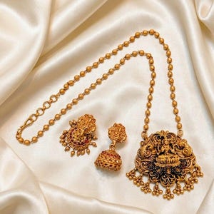 May include: A gold-toned jewelry set consisting of a necklace and a pair of jhumka earrings. The necklace features a beaded chain and a large, ornate pendant with a central deity figure. The earrings match the pendant, featuring red accents.