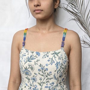 May include: A cream-colored dress featuring a blue and green floral print, a square neckline, and adjustable straps decorated with small, vibrant rainbow-colored beads and purple metal sliders, creating a distinct and colorful visual detail.