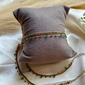 Könnte beinhalten: Goldfarbene Fußkettchen mit smaragdgrünen Akzenten, präsentiert auf einem grauen Kissen. Die Fußkettchen haben zarte Ketten und kleine, tropfenförmige grüne Steine. Der Schmuck wird auf einer weißen Satin-Oberfläche präsentiert.