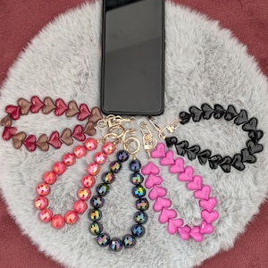 Könnte beinhalten: Fünf dekorative Handy-Handschlaufen, die um ein schwarzes Smartphone angeordnet sind. Die Schlaufen bestehen aus verschiedenen Perlenformen, darunter rote, rosa und schwarze Herzen sowie irisierende und facettierte Rundperlen, befestigt mit goldfarbenen Metallverschlüssen.
