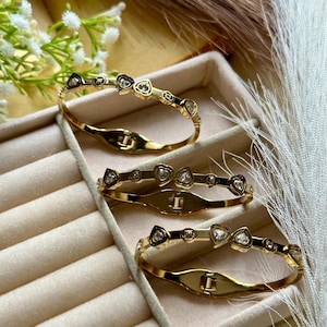 Könnte beinhalten: Goldfarbene Armbänder mit herzförmigen und runden, klaren Edelsteinen. Die Armbänder werden in einer beigen Schmuckschatulle präsentiert. Die Armbänder haben einen Verschluss. Modeschmuck.