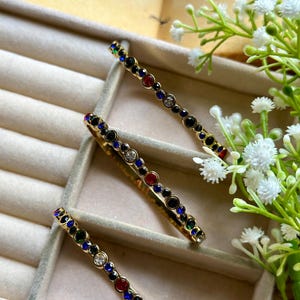 Könnte beinhalten: Drei goldfarbene Haarspangen mit einer Reihe farbiger Edelsteine. Die Edelsteine umfassen rote, blaue, grüne, schwarze und klare Kristalle. Die Spangen sind in einer samtgefütterten Schmuckschatulle mit weißen Blumen im Hintergrund angeordnet.