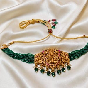 Può includere: Una collana girocollo color oro con un ciondolo centrale ornato da figure in rilievo, accentuato da piccole pietre rosse e verdi. La collana è composta da più fili di minuscole perline verdi e include pendenti con perline verdi e perle bianche.