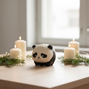 May include: A wooden panda figurine with black and white details sits on a wooden table, flanked by lit white candles and greenery. The panda has a carved appearance. The scene is illuminated by natural light, creating a cozy ambiance.