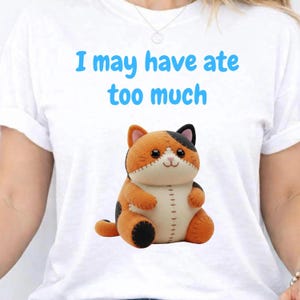 May include: A white t-shirt featuring the blue text I may have ate too much above a graphic of a round, plush-style calico cat with orange, black, and cream-colored patches, stitched details, and a small, smiling face with dark eyes.