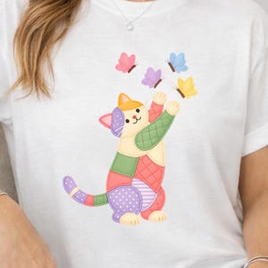 May include: A white t-shirt featuring a graphic of a patchwork-style cat with various coloured segments including green, pink, purple, and orange. The cat is reaching up toward three small butterflies in pink, purple, and yellow colours.