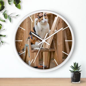 Puede incluir: Reloj de pared redondo con marco blanco. La imagen muestra un gato calicó con casco de construcción y cinturón de herramientas, trabajando en una estructura de madera. El gato está en una pequeña escalera de madera. Las manecillas son blancas.
