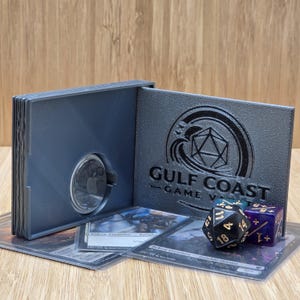 Puede incluir: Un juego de cartas y dados grises con el texto "GULF COAST GAME VAULT". El juego incluye un tarjetero gris, dos dados y varias cartas de juego. Los dados son negros y morados.