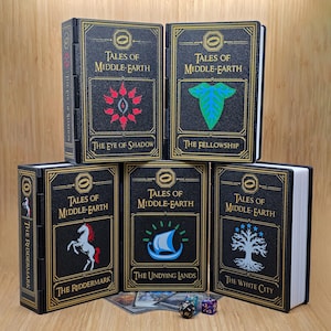 Könnte beinhalten: Fünf schwarze Aufbewahrungsboxen mit goldenen Verzierungen und dem Text "Tales of Middle-Earth". Jede Box zeigt ein anderes Design und einen anderen Titel: "The Eye of Shadow", "The Fellowship", "The Riddermark", "The Undying Lands" und "The White City".