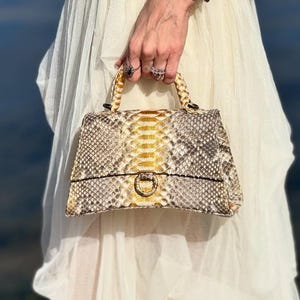 May include: A snakeskin handbag with a gold clasp and handle is held up. The bag features a mix of tan, brown, and gold tones. A silver watch with a blue face is worn on the wrist.