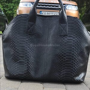 Op de afbeelding: Zwarte leren handtas met een getextureerd, schubachtig patroon. De tas heeft een afgeronde vorm, een handvat aan de bovenkant en een ritssluiting. De merknaam "RoyalHouseAtelier" is zichtbaar. Het ontwerp suggereert een slangenhuideffect.