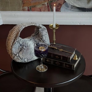 Könnte beinhalten: Eine Handtasche mit Schlangenmuster steht auf einem dunklen, runden Tisch neben einem Cocktail in einem Stielglas. Eine brennende Kerze in einem goldenen Halter, zwei Bücher und eine kleine Glocke befinden sich ebenfalls auf dem Tisch. Der Hintergrund zeigt ein Wandgemälde.