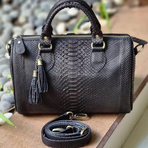May include: Black leather handbag with a snakeskin pattern, featuring a top handle and a detachable shoulder strap. The bag has a zipper closure and gold-colored hardware, including a tassel charm. The bag is approximately 10 inches wide.