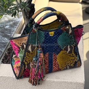 Puede incluir: Un bolso colorido con un estampado de piel de serpiente. El bolso presenta una variedad de colores, incluyendo azul, verde, amarillo, rosa y marrón. Tiene dos asas y un borla decorativa. El bolso está sobre una superficie blanca.