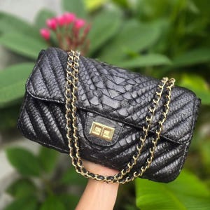 Pode incluir: Bolsa de mão de couro preto acolchoada com corrente e fecho dourados. A bolsa tem um fecho de aba e um padrão texturizado semelhante à pele de cobra. A corrente dourada é drapeada sobre a bolsa. O fundo está desfocado com folhagem verde e flores cor-de-rosa.