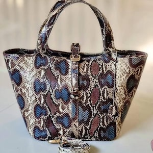 Può includere: Una borsa tote con motivo a pelle di serpente con manico superiore e tracolla staccabile. La borsa presenta un mix di colori blu, viola e crema. Ha una chiusura e finiture color oro.