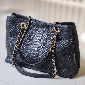 Op de afbeelding: Zwarte tote bag met een getextureerd, reptielachtig patroon. De tas heeft goudkleurige kettingriemen en hardware. Het ontwerp omvat een handvat aan de bovenkant en een schouderriem. Het materiaal van de tas lijkt leer te zijn.