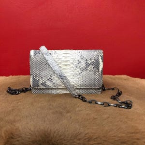 Pode incluir: Uma clutch retangular com um padrão de pele de cobra em branco, cinza e preto, com uma alça a condizer e uma corrente escura. A bolsa está colocada sobre um fundo vermelho e uma superfície marrom e peluda.