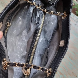 K&ouml;nnte beinhalten: Schwarze Handtasche mit goldfarbenen Kettenriemen und Rei&szlig;verschluss. Der Innenraum ist mit grauem Stoff gef&uuml;ttert und enth&auml;lt eine Rei&szlig;verschlusstasche. Die Tasche besteht aus einem strukturierten Material, m&ouml;glicherweise Leder oder leder&auml;hnlichem Material.