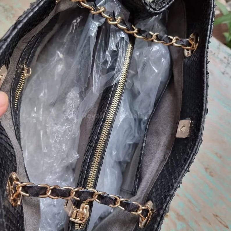 K&ouml;nnte beinhalten: Schwarze Handtasche mit goldfarbenen Kettenriemen und Rei&szlig;verschluss. Der Innenraum ist mit grauem Stoff gef&uuml;ttert und enth&auml;lt eine Rei&szlig;verschlusstasche. Die Tasche besteht aus einem strukturierten Material, m&ouml;glicherweise Leder oder leder&auml;hnlichem Material.