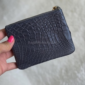 Pode incluir: Uma carteira cinza escuro com um padrão texturizado semelhante à pele de réptil. A carteira pequena e quadrada tem um zíper dourado e um puxador correspondente. A carteira está sobre um fundo branco e fofo.