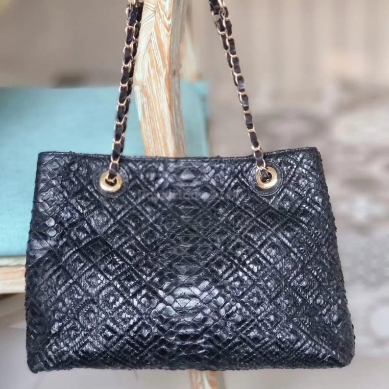K&ouml;nnte beinhalten: Schwarze, gesteppte Leder-Tote-Tasche mit goldenen Kettenriemen. Die Tasche hat ein Rautenmuster und goldfarbene Beschl&auml;ge. Die Kettenriemen sind mit goldenen Ringen an der Tasche befestigt. Die Tasche liegt auf einer hellen Holzoberfl&auml;che.