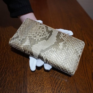 Op de afbeelding: Een rechthoekige portemonnee met een slangenpatroon in tinten beige, bruin en grijs. De portemonnee heeft een goudkleurige ritssluiting. Het item wordt vastgehouden door een persoon met witte handschoenen.