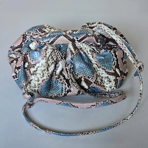 May include: A round handbag with a snakeskin pattern in shades of blue, pink, and brown. The bag has a knotted top and a long, thin strap. The overall design is soft and textured.