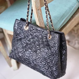 K&ouml;nnte beinhalten: Schwarze Tragetasche mit strukturierter, rautenf&ouml;rmiger Au&szlig;enseite. Die Tasche hat goldfarbene Kettenriemen und Beschl&auml;ge. Die Tasche h&auml;ngt an einem Holzstuhl.