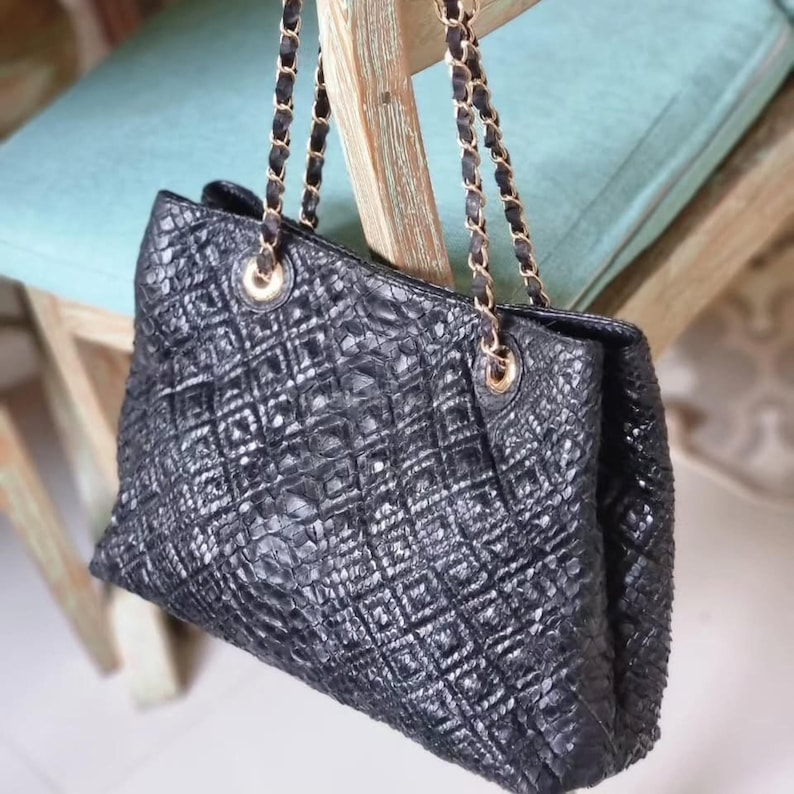 K&ouml;nnte beinhalten: Schwarze Tragetasche mit strukturierter, rautenf&ouml;rmiger Au&szlig;enseite. Die Tasche hat goldfarbene Kettenriemen und Beschl&auml;ge. Die Tasche h&auml;ngt an einem Holzstuhl.