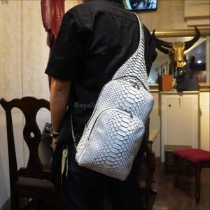 Pode incluir: Bolsa de ombro branca e cinza com padrão de pele de cobra e uma única alça de ombro. A bolsa tem um compartimento principal com zíper e um bolso frontal menor. A bolsa é usada no ombro.