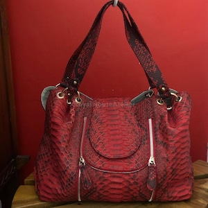 Può includere: Una borsa rossa con un motivo a pelle di serpente. La borsa ha due cerniere sul davanti, una chiusura a patta e due manici. I manici e la parte superiore della borsa sono di una tonalità di rosso più scura.