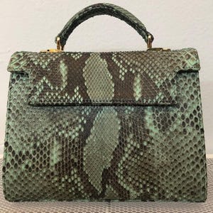 Puede incluir: Bolso estructurado con asa superior y cierre de solapa. El bolso presenta un estampado de piel de serpiente en tonos verdes y marrones, con herrajes dorados. El asa también es de piel de serpiente.