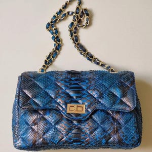 Pode incluir: Bolsa acolchoada de pele de cobra azul e preta com fecho dourado. A bolsa apresenta uma alça de corrente com elos dourados e azuis. A bolsa é retangular com fecho de aba.