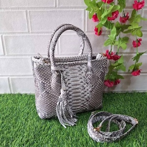 Pode incluir: Uma mala cinza e branca com padrão de pele de cobra, com duas alças e uma alça de ombro removível. A mala tem uma borla e está sobre um leito de relva verde. A mala tem cerca de 25 cm de largura.