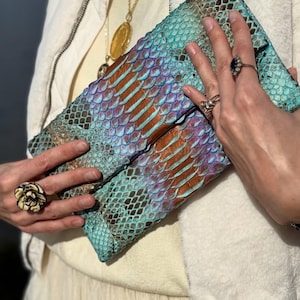 Può includere: Una pochette con motivo a pelle di serpente turchese, marrone e viola. La pochette è aperta, rivelando l'interno. La borsa ha un design pieghevole. La tavolozza dei colori comprende sfumature di blu, marrone e viola.