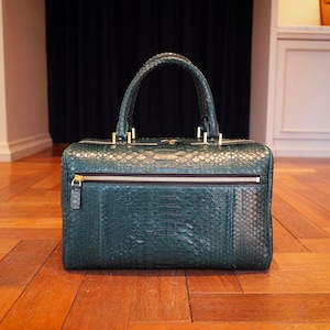 Puede incluir: Bolso de mano rectangular verde oscuro con un patrón texturizado similar a la piel de serpiente. El bolso tiene un asa superior, un bolsillo frontal con cremallera y herrajes dorados. El bolso está sobre un suelo de madera.