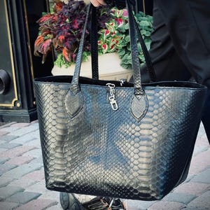Puede incluir: Bolso tote negro con estampado de piel de serpiente. El bolso tiene dos asas y un cierre plateado. El bolso se sostiene frente a un fondo de flores y un edificio.