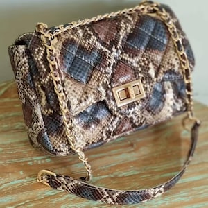 Pode incluir: Bolsa acolchoada com padrão de pele de cobra em tons de castanho, bege e preto. A bolsa apresenta uma alça de corrente dourada e um fecho retangular. O design da bolsa inclui um padrão de losangos.
