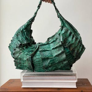 May include: A green, textured handbag with a shoulder strap. The bag is made of a scaly material, possibly leather or a similar fabric, and has a unique, ruffled design. The bag is displayed on a clear acrylic box.