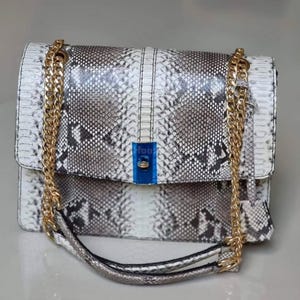 Pode incluir: Bolsa com padrão de pele de cobra branca, cinza e preta, com uma alça de corrente dourada. A bolsa retangular tem um fecho de aba e um fecho dourado. A alça é uma corrente dourada. A bolsa é de cor neutra.