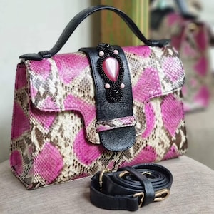 Pode incluir: Bolsa com padrão de pele de cobra rosa, castanho e creme, com alça e alça pretas. O fecho da aba apresenta uma alça de couro preta com uma pedra preciosa rosa e enfeites de contas. Inclui uma alça de ombro preta removível com ferragens douradas.