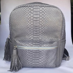 May include: Gray backpack with a snakeskin pattern. The bag has a front zippered pocket, side pockets, and two decorative tassels. The backpack has adjustable shoulder straps and a top handle.