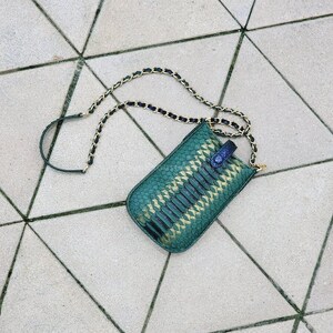 May include: A teal green snakeskin-patterned phone bag with a gold and black chain strap. The bag has a rectangular shape with a rounded top and a small blue flap closure. The bag is lying on a light grey tiled surface.