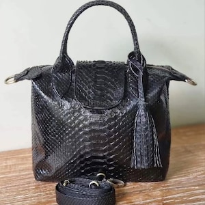 Pode incluir: Bolsa de mão de couro preta com um padrão de pele de cobra texturizado. A bolsa possui uma alça superior, um fecho de aba e uma alça de ombro removível. Uma borla decorativa pende da lateral. A bolsa está sobre uma superfície de madeira.