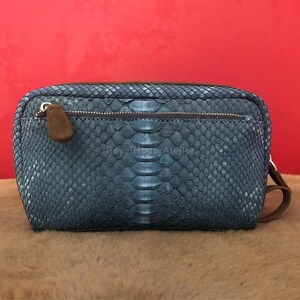 Puede incluir: Un clutch rectangular de piel de serpiente azul con cierre de cremallera y tirador de ante marrón. El clutch tiene un ribete de cuero marrón y una correa de muñeca de ante marrón. El fondo es rojo liso.