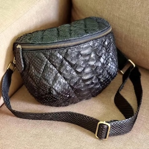 Può includere: Marsupio nero con un design trapuntato e strutturato. La borsa presenta una chiusura con cerniera e una tracolla regolabile con finiture metalliche color oro. La borsa è realizzata in un materiale che ricorda la pelle di serpente.