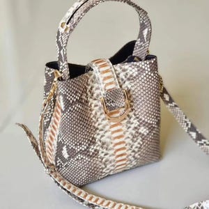 May include: A small bucket bag with a snakeskin pattern in grey, brown, and cream. It has a top handle, a detachable shoulder strap, and a gold-coloured buckle. The interior of the bag is black.